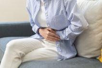 žena u plavoj košulji sedi na kauču i drži obe ruke položene na levu stranu stomaka