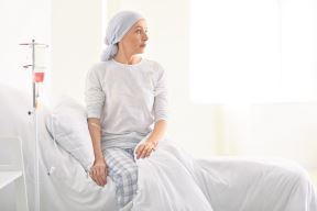 Žena sedi na krevetu, s turbanom na glavi, prima hemoterapiju – bori se protiv raka.