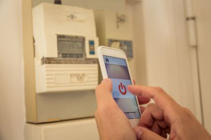 čovek proverava potrošnju struje na mobilnom telefonu dok se u pozadini vidi pametno brojilo