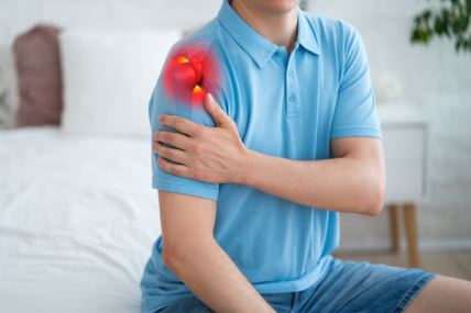 muškarac u plavoj majci sedi na ivisi kreveta i drži levom rukom desno rame koje ga boli