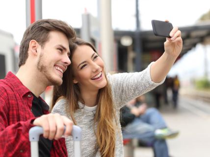 Par putnika koji fotografiše selfi pametnim telefonom na železničkoj stanici