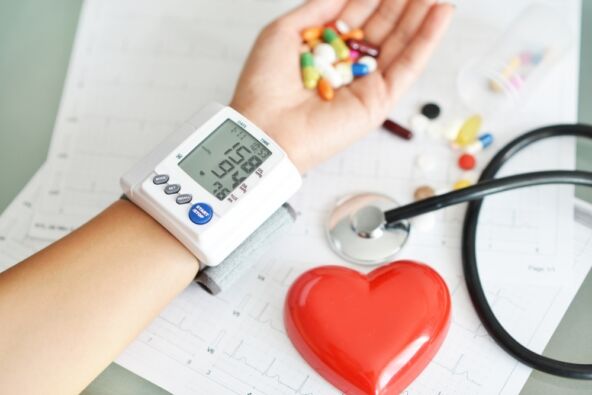 Žena sa hipertenzijom meri krvni pritisak i u šaci drži mnoštvo lekova, EKG nalazi na stolu i model srca