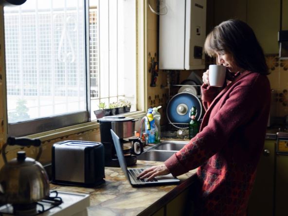 Žena stoji u kuhinji, ispija kafu i radi na laptopu